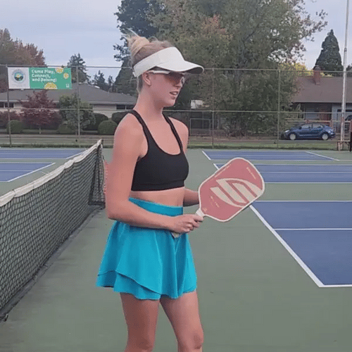 A woman on a pickleball court wearing a black sports bra, a bright turquoise tennis skirt, and a white visor, holding a paddle 